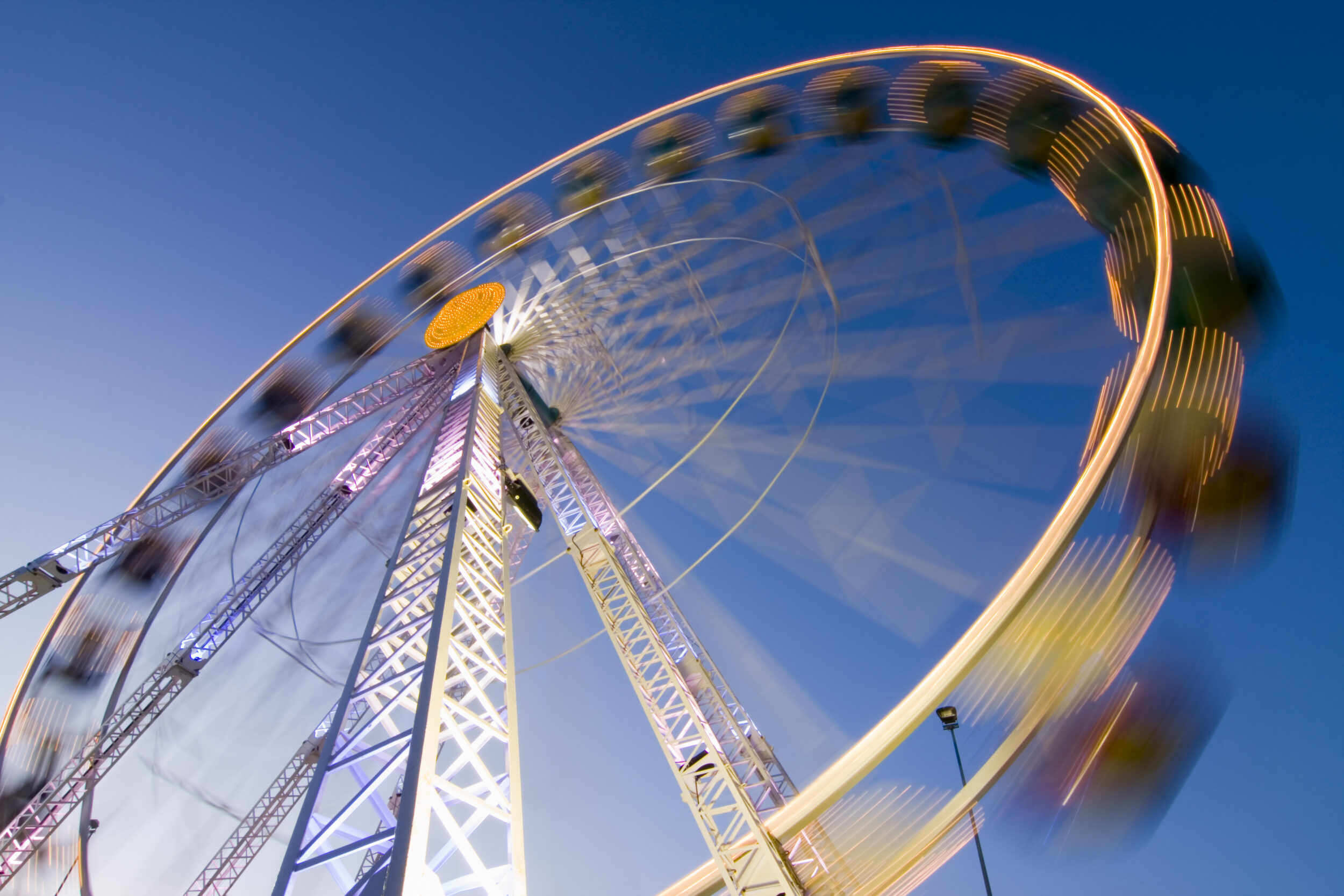 Ein Riesenrad dreht sich vor blauem Himmel. Durch eine Langzeitbelichtung entstehen leuchtende Lichtspuren, die die Bewegung sichtbar machen.