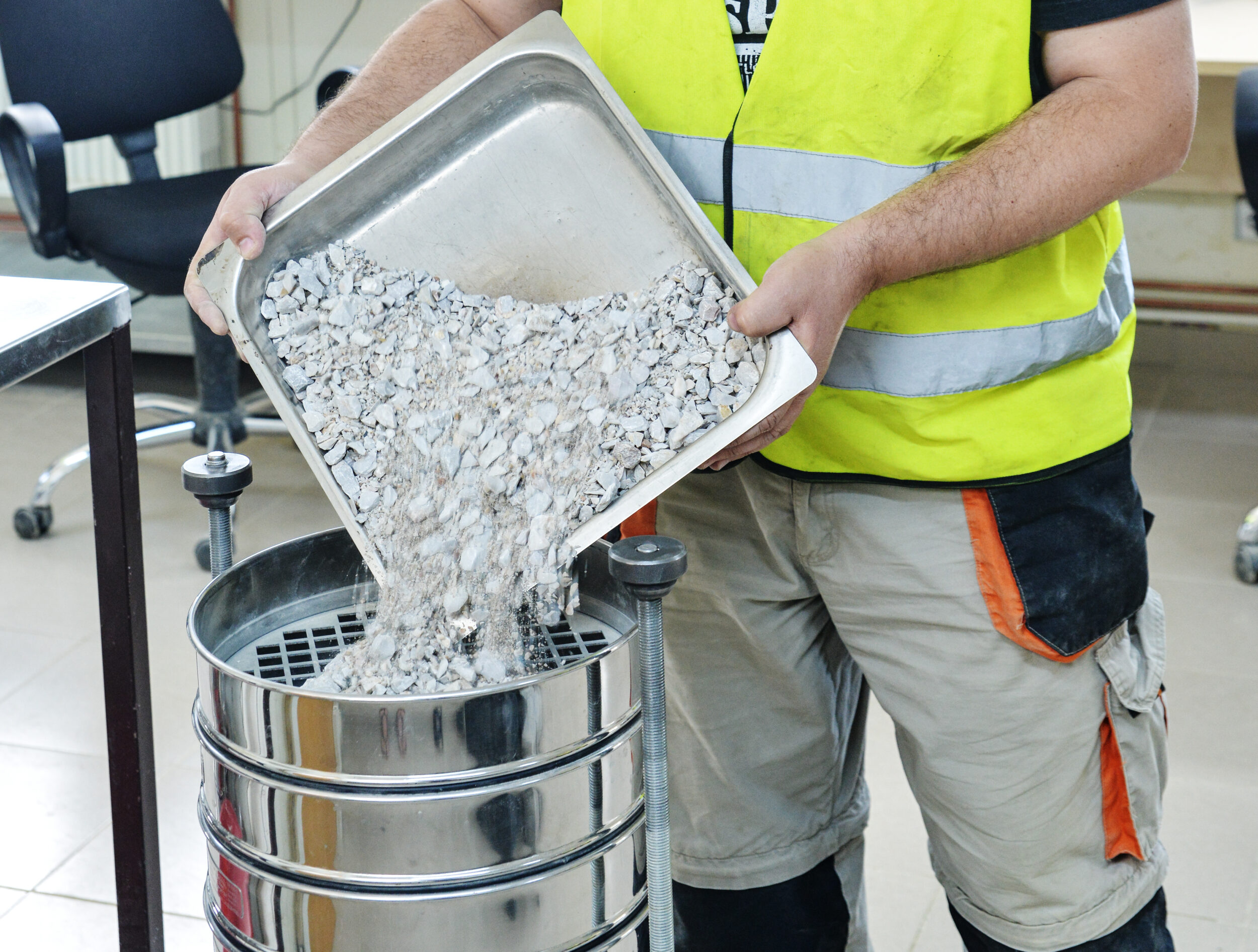 Das Bild zeigt eine Person mit gelber Sicherheitsweste und beigefarbener Hose, die in einem Labor- oder Industriebereich zerkleinerte Steine oder Kies aus einem Metallbehälter in ein Siebgerät schüttet. Die Siebe sind übereinandergestapelt und dienen vermutlich der Materialanalyse.