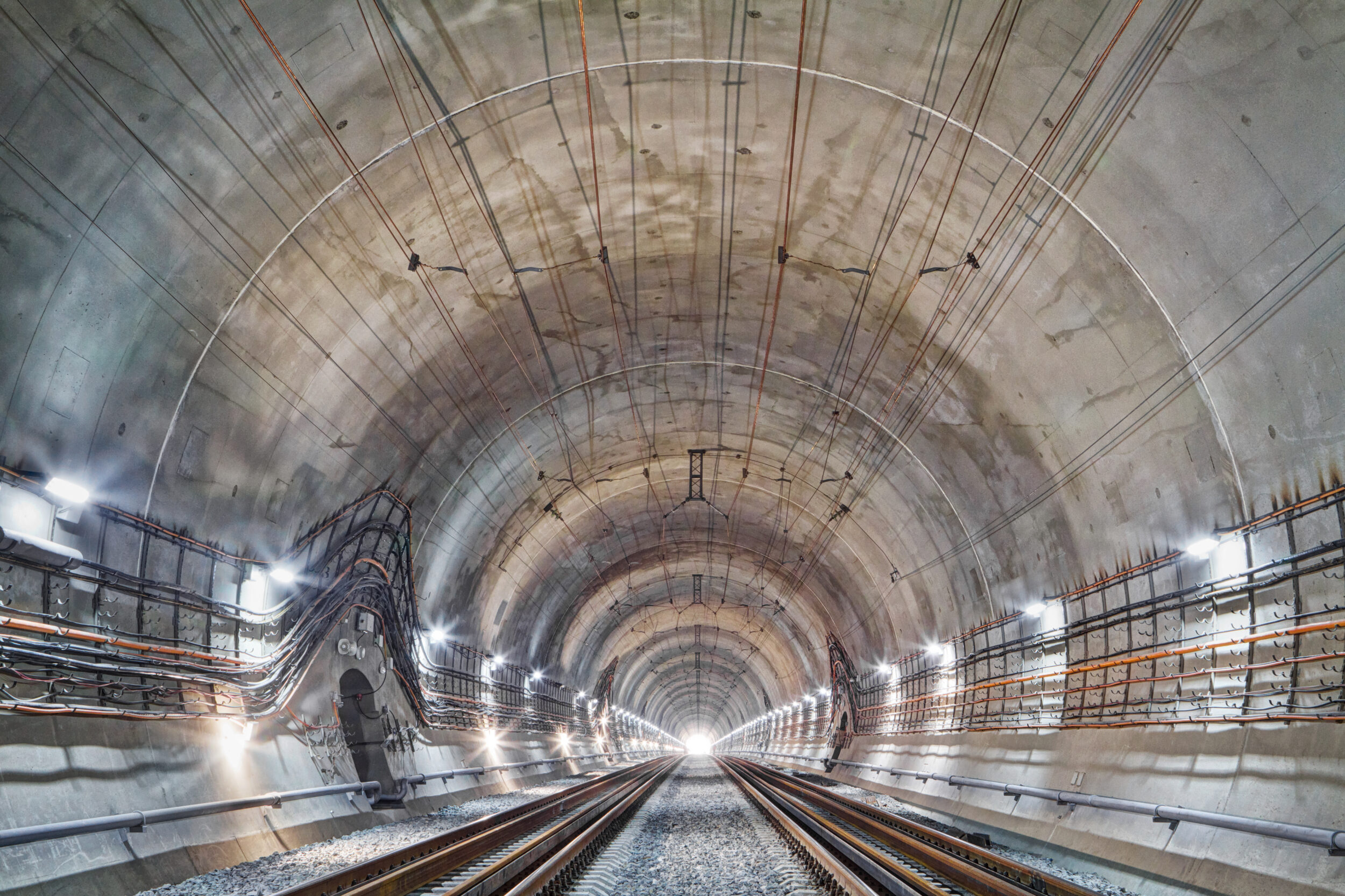 Das Bild zeigt das Innere eines großen, gut beleuchteten Tunnels mit zwei Gleisen. Entlang der Tunnelwände verlaufen verschiedene Kabel und Rohre, und auf beiden Seiten sind regelmäßig Lampen angebracht. Der Blick führt in Richtung eines hellen Lichtpunkts am Ende des Tunnels.