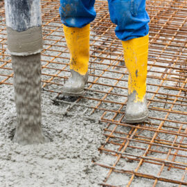 Das Bild zeigt einen Bauarbeiter mit gelben Gummistiefeln und blauer Hose, der auf einem Bewehrungsgitter steht. Aus einem Schlauch wird frischer Beton auf das Gitter gegossen, um eine Betondecke im Rohbau herzustellen. Die Szene zeigt einen typischen Arbeitsschritt beim Betonieren einer Geschossdecke.