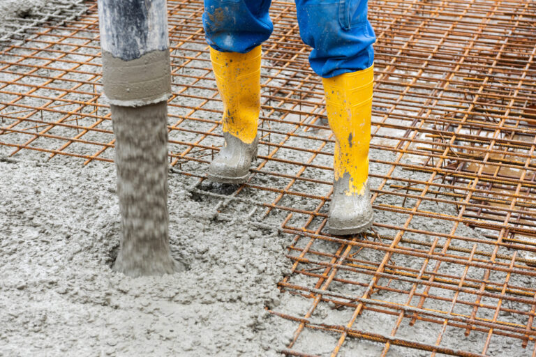 Das Bild zeigt einen Bauarbeiter mit gelben Gummistiefeln und blauer Hose, der auf einem Bewehrungsgitter steht. Aus einem Schlauch wird frischer Beton auf das Gitter gegossen, um eine Betondecke im Rohbau herzustellen. Die Szene zeigt einen typischen Arbeitsschritt beim Betonieren einer Geschossdecke.