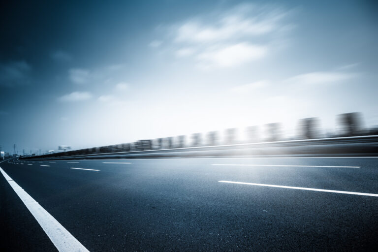 Das Bild zeigt einen Abschnitt einer Asphaltstraße mit deutlich sichtbaren Fahrbahnmarkierungen. Weiße gestrichelte Linien trennen die Fahrspuren, und am linken Rand verläuft eine durchgezogene weiße Linie. Im Hintergrund sind Bauwerke und ein teilweise bewölkter Himmel zu erkennen.