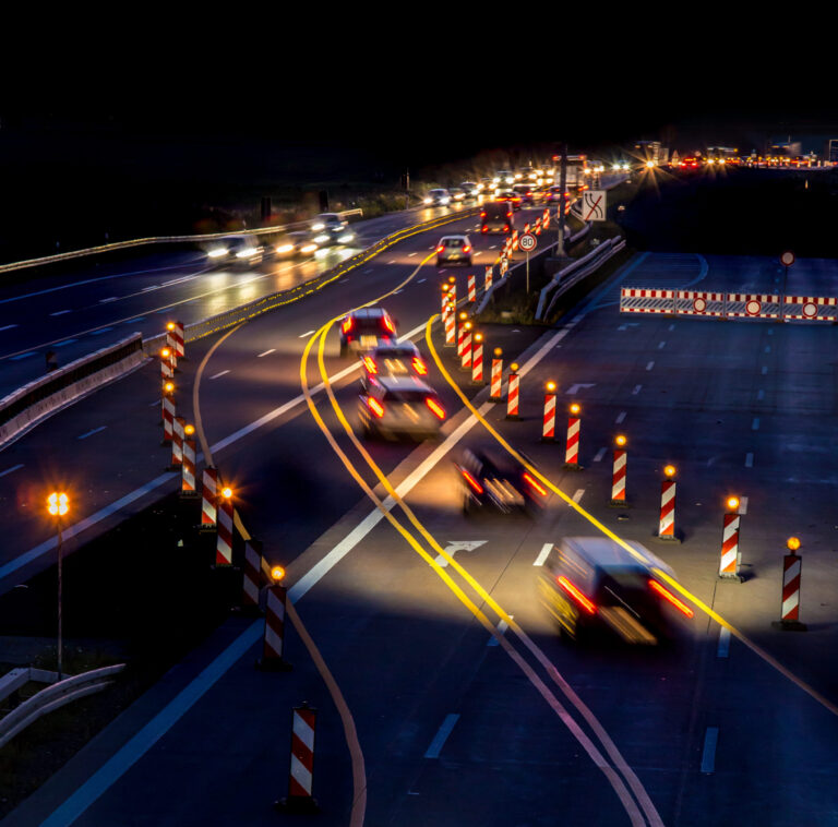 Das Bild zeigt eine nächtliche Szene auf einer mehrspurigen Autobahn mit mehreren fahrenden Autos. Durch die Langzeitbelichtung entstehen Lichtspuren von Scheinwerfern und Rücklichtern. Zahlreiche Verkehrsleitkegel und Absperrungen mit reflektierenden Streifen deuten auf Bauarbeiten hin.