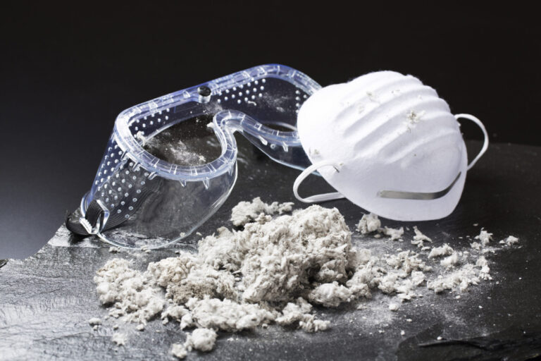 Das Bild zeigt eine Schutzbrille und eine weiße Staubmaske, die auf einer dunklen Oberfläche liegen. Davor befindet sich ein Haufen faseriges Material, das wie Asbest oder ein ähnlicher Stoff aussieht. Der schwarze Hintergrund hebt die Gegenstände im Vordergrund deutlich hervor.
