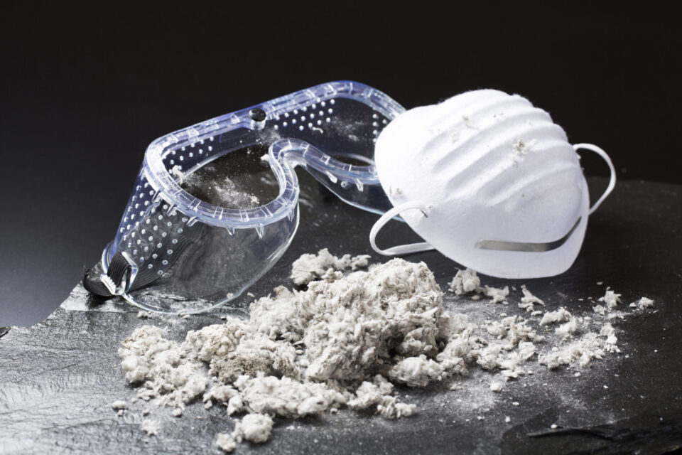 Das Bild zeigt eine Schutzbrille und eine weiße Staubmaske, die auf einer dunklen Oberfläche liegen. Davor befindet sich ein Haufen faseriges Material, das wie Asbest oder ein ähnlicher Stoff aussieht. Der schwarze Hintergrund hebt die Gegenstände im Vordergrund deutlich hervor.