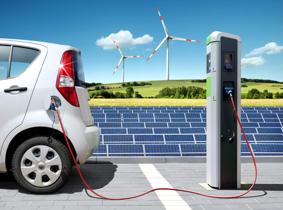 Das Bild zeigt ein Elektroauto, das an einer Ladestation mit einem roten Kabel angeschlossen ist. Im Hintergrund befinden sich Solarpaneele und Windräder, die auf die Nutzung erneuerbarer Energien hinweisen. Die Szene spielt sich im Freien an einem sonnigen Tag mit blauem Himmel und einigen Wolken ab.