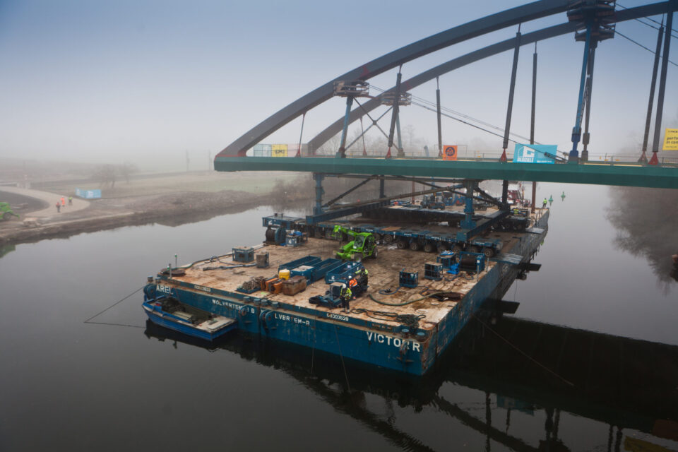 Das Bild zeigt ein großes Bauschiff unter einer Brücke, ausgestattet mit verschiedenen Baumaschinen und Kränen. Auf der Brücke darüber befinden sich weitere Geräte und Arbeiter, was auf Bau- oder Wartungsarbeiten hinweist. Die Umgebung ist neblig, wodurch die Sicht auf die Landschaft eingeschränkt ist.