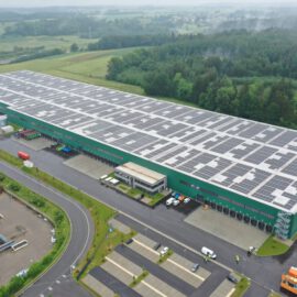 Das Bild zeigt ein großes Industriegebäude mit grüner Fassade und zahlreichen Solarpanels auf dem Dach, umgeben von Grünflächen, kleineren Gebäuden, Parkplätzen, Straßen sowie Feldern und Wäldern in ländlicher Umgebung.