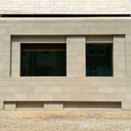 Das Bild zeigt die Außenfassade eines Gebäudes mit beigefarbenen rechteckigen Natursteinplatten, die horizontal angeordnet sind. Zwei große rechteckige Fenster mit dunklem Glas spiegeln die Umgebung. Der Boden vor dem Gebäude besteht aus Erde oder Kies.