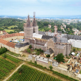 Das Bild zeigt eine Luftaufnahme eines großen historischen Gebäudekomplexes, der sich in Renovierung befindet. Mehrere Türme und Gebäudeteile sind eingerüstet, im Vordergrund sind Weinberge zu sehen, rechts ein Parkbereich. Im Hintergrund erstreckt sich eine Stadtlandschaft mit Wohnhäusern und weiteren Gebäuden.