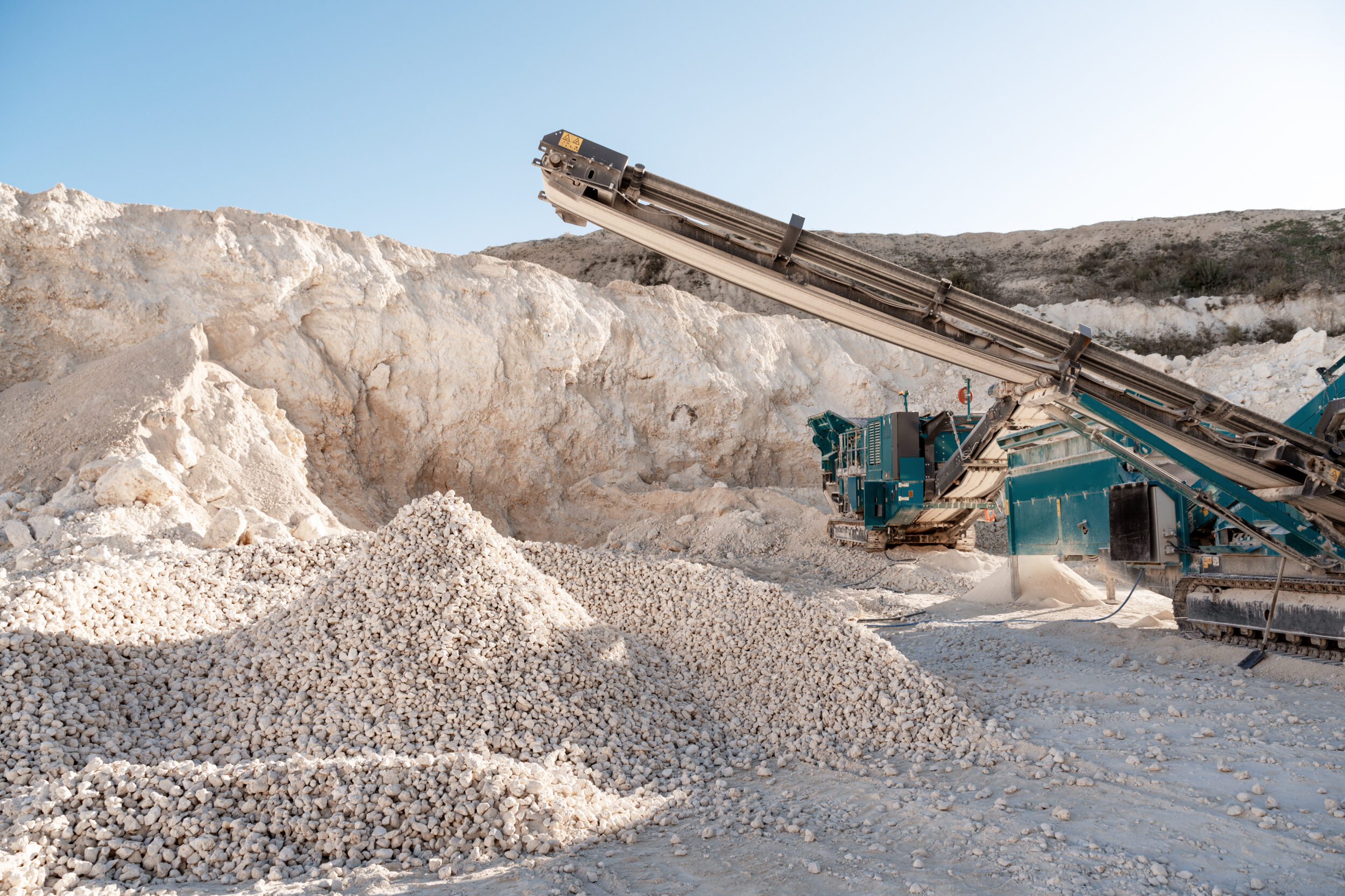 Das Bild zeigt eine große Maschine mit Förderband, die in einem Steinbruch arbeitet. Vor der Maschine liegen mehrere Haufen heller, grober Steine, während im Hintergrund eine hohe Felswand zu sehen ist. Die Szene ist von hellem Sonnenlicht beleuchtet und vermittelt den Eindruck intensiver Materialverarbeitung.