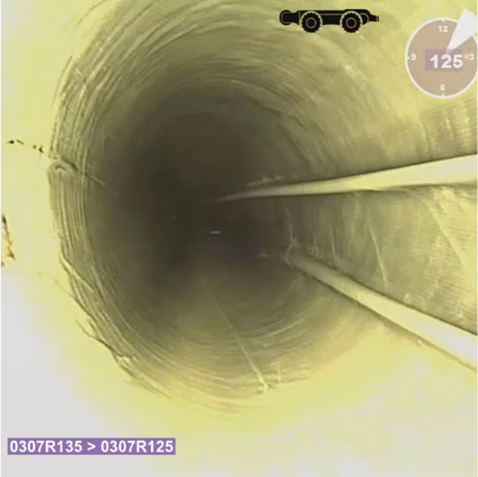 Das Bild zeigt die Innenansicht eines runden Rohres, aufgenommen mit einer Inspektionskamera. Die Rohrwand ist gleichmäßig strukturiert und verläuft in die Tiefe, wodurch ein Tunnel-Effekt entsteht. Auf der rechten Seite sind zwei parallel verlaufende Leitungen oder Kabel sichtbar, und oben links befindet sich eine grafische Anzeige. Unten links ist ein kleiner Textbereich mit Zahlen und Buchstaben eingeblendet, der vermutlich die Position oder den Abschnitt des Rohres angibt.