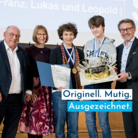 Auf dem Bild steht eine Gruppe von f&uuml;nf Personen auf einer B&uuml;hne und blickt l&auml;chelnd in die Kamera. Zwei Jugendliche in der Mitte tragen Medaillen, einer h&auml;lt eine Urkunde und der andere ein Modell, w&auml;hrend die Erwachsenen danebenstehen. Im Vordergrund befindet sich ein gro&szlig;er Schriftzug, der auf eine Auszeichnung hinweist.