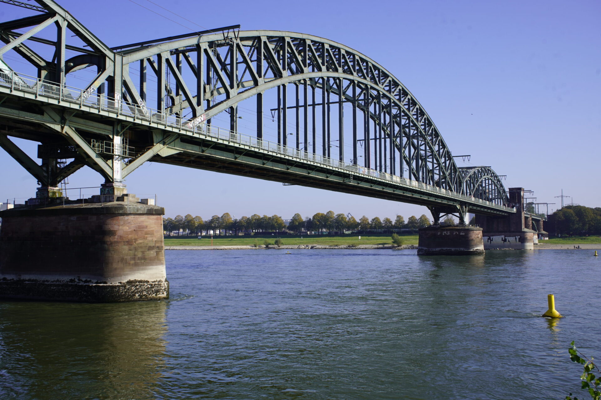 Auf dem Bild ist eine große Stahlbrücke mit mehreren Bögen zu sehen, die über einen breiten Fluss führt. Die Brücke ruht auf massiven Pfeilern und verbindet zwei Ufer mit einer flachen Landschaft im Hintergrund. Das Wasser fließt ruhig unter der Konstruktion hindurch und spiegelt Teile der Brücke wider.