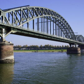 Auf dem Bild ist eine gro&szlig;e Stahlbr&uuml;cke mit mehreren B&ouml;gen zu sehen, die &uuml;ber einen breiten Fluss f&uuml;hrt. Die Br&uuml;cke ruht auf massiven Pfeilern und verbindet zwei Ufer mit einer flachen Landschaft im Hintergrund. Das Wasser flie&szlig;t ruhig unter der Konstruktion hindurch und spiegelt Teile der Br&uuml;cke wider.