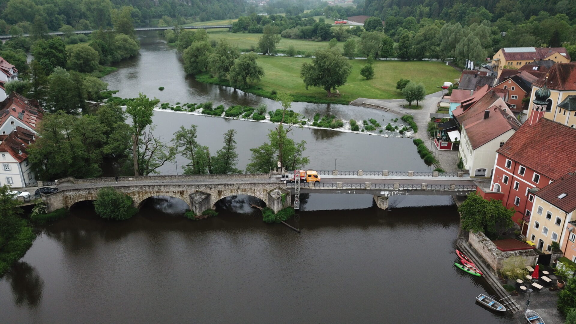Eine Luftaufnahme zeigt eine steinerne Brücke, die einen Fluss in einer kleinen Ortschaft überquert. Auf der Brücke steht ein Baustellenfahrzeug neben einem Brückenuntersichtgerät, das für Arbeiten oder Prüfungen an der Brücke eingesetzt wird. Im Hintergrund sind ein Wehr, weitläufige Grünflächen, Bäume und mehrere Gebäude entlang des Flussufers zu sehen.