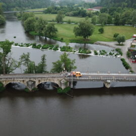 Eine Luftaufnahme zeigt eine steinerne Brücke, die einen Fluss in einer kleinen Ortschaft überquert. Auf der Brücke steht ein Baustellenfahrzeug neben einem Brückenuntersichtgerät, das für Arbeiten oder Prüfungen an der Brücke eingesetzt wird. Im Hintergrund sind ein Wehr, weitläufige Grünflächen, Bäume und mehrere Gebäude entlang des Flussufers zu sehen.