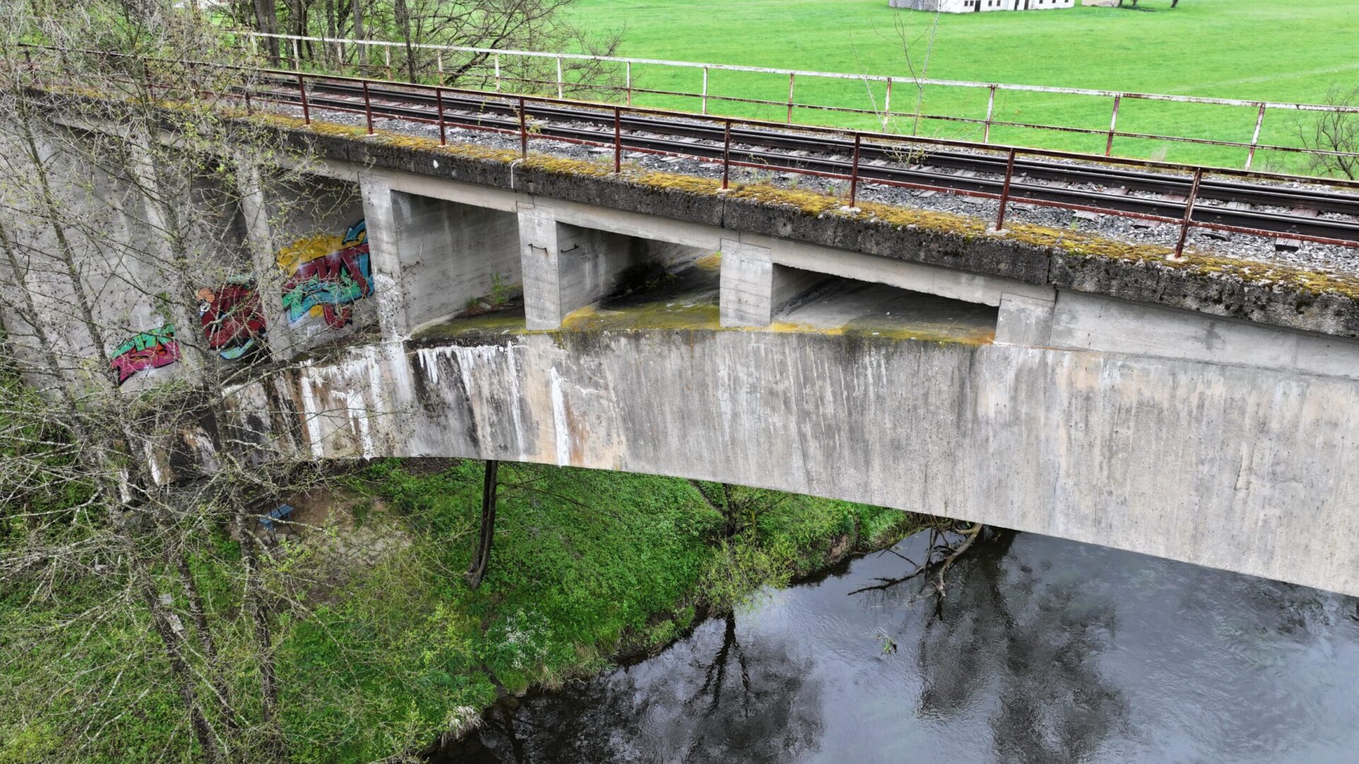 Die Aufnahme zeigt eine Beton-Eisenbahnbrücke aus der Nähe, die einen Fluss überquert. Auf der Brücke verlaufen Gleise mit einem Geländer, während unterhalb das Flussufer sowie die Brückenkonstruktion mit mehreren Stützen und Öffnungen zu sehen sind.