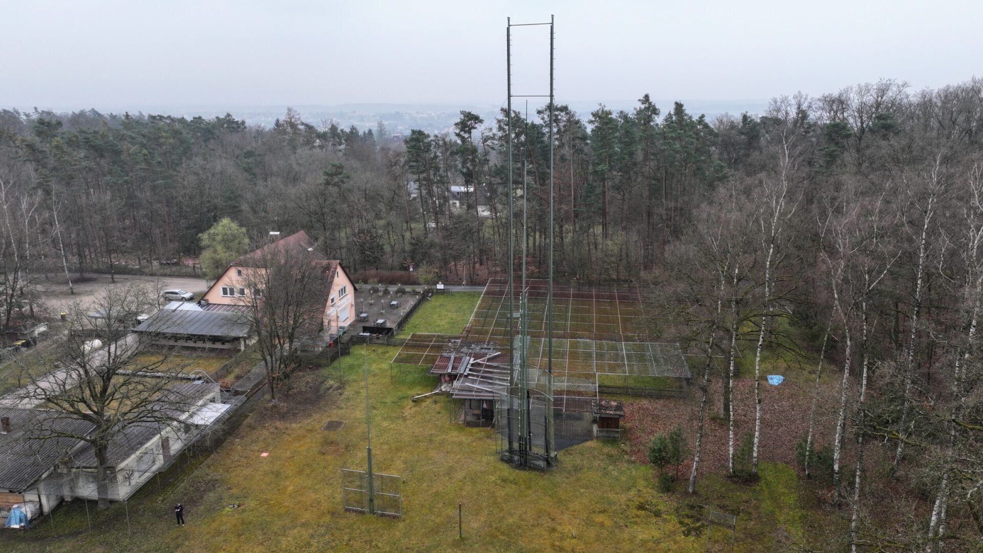 Die Luftaufnahme zeigt eine Sportanlage mit mehreren Tennisplätzen, umgeben von Bäumen und Grünflächen. Im Vordergrund sind hohe Ballfangzäune zu sehen, während sich im Hintergrund ein Vereinsgebäude und ein Waldgebiet erstrecken.