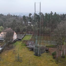 Die Luftaufnahme zeigt eine Sportanlage mit mehreren Tennisplätzen, umgeben von Bäumen und Grünflächen. Im Vordergrund sind hohe Ballfangzäune zu sehen, während sich im Hintergrund ein Vereinsgebäude und ein Waldgebiet erstrecken.