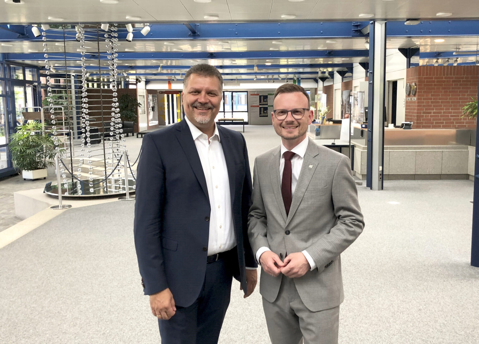 Zwei Personen stehen in einem modernen, großzügigen Innenraum mit hoher Decke und großen Fenstern; im Hintergrund sind Schreibtische, Pflanzen und eine dekorative Struktur aus hängenden Kugeln zu sehen, beide tragen formelle Kleidung.