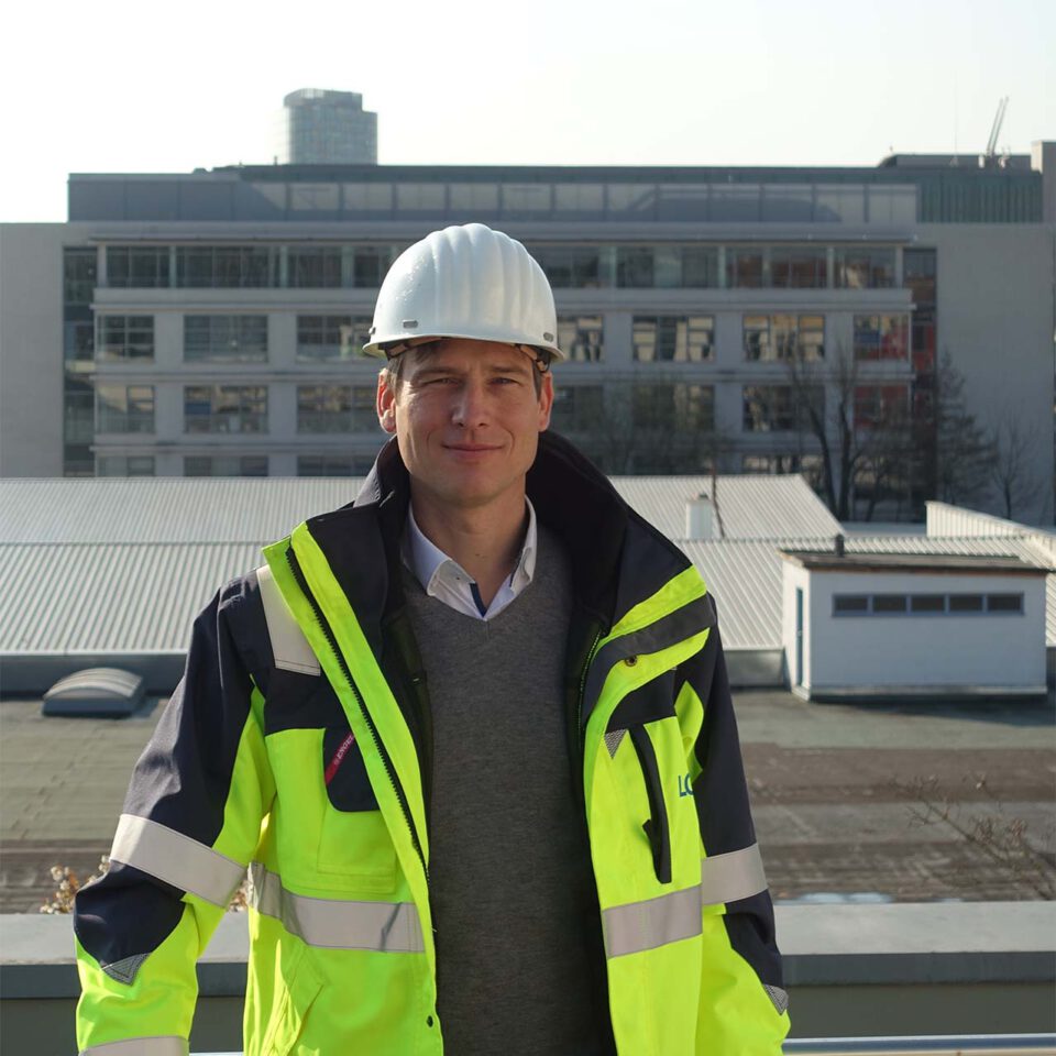 Eine Person mit weißem Schutzhelm und Warnweste steht vor einem Industrie- oder Bürogebäude.