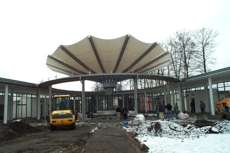Baustelle mit großer, runder Struktur und zeltartigem Dach auf Stützen; mehrere Bauarbeiter und ein gelber Bagger; schneebedeckter Boden mit Erdhaufen und Baumaterialien, im Hintergrund kahle Bäume.