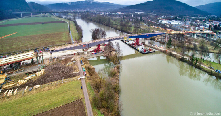 Brückenbaustelle über einem Fluss in ländlicher Umgebung, mit sichtbaren Baumaschinen und Materialien auf beiden Uferseiten; umgeben von Feldern, Straßen, Hügeln und einer Ortschaft im Hintergrund, aufgenommen aus erhöhter Perspektive.