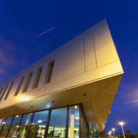 Modernes Gebäude mit kantigem Design, großen Glasfenstern im unteren Bereich und glatter, hellfarbener Fassade mit vertikalen Fensterschlitzen im oberen Bereich; aufgenommen in der Dämmerung mit Straßenbeleuchtung und tiefblauem Himmel im Hintergrund.