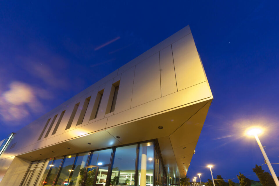 Modernes Gebäude mit kantigem Design, großen Glasfenstern im unteren Bereich und glatter, hellfarbener Fassade mit vertikalen Fensterschlitzen im oberen Bereich; aufgenommen in der Dämmerung mit Straßenbeleuchtung und tiefblauem Himmel im Hintergrund.