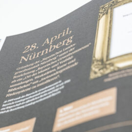 Das Bild zeigt ein aufgeklapptes Buch mit einer Doppelseite. Auf der linken Seite befindet sich ein Text zur Gründung der Bayerischen Landesgewerbeanstalt in Nürnberg, auf der rechten Seite ein gerahmtes historisches Dokument.