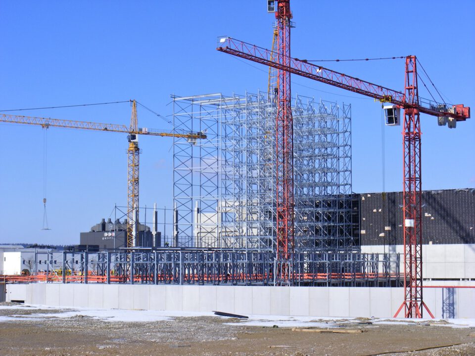 Eine Baustelle mit drei Kränen – zwei roten und einem gelben – sowie einem im frühen Bau befindlichen Stahlgerüst; im Hintergrund sind weitere Industriegebäude und Geräte zu sehen, der schneebedeckte Boden deutet auf winterliche Bedingungen hin.