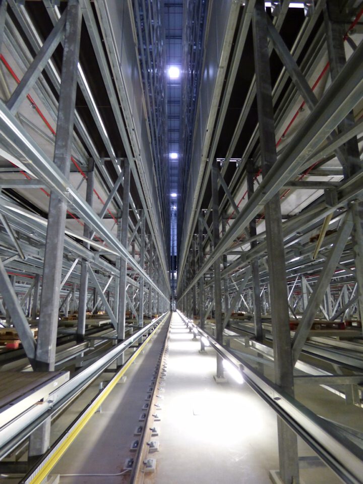 Ein langer, schmaler Gang in einem Industrie- oder Lagergebäude, gesäumt von hohen Metallregalen mit symmetrischem Trägergerüst; Bodenleuchten beleuchten den Weg, und die Decke mit weiterer Beleuchtung ist in großer Höhe sichtbar.