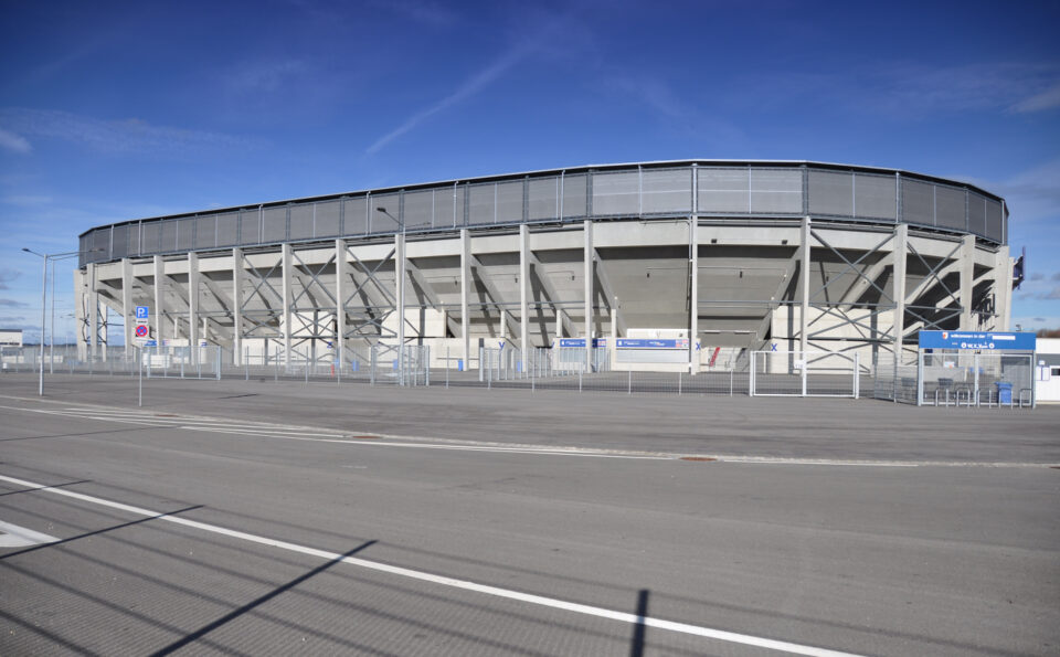 Außenansicht eines großen, modernen Stadions mit kreisförmiger Struktur, mehreren Stützträgern und umlaufendem Metallzaun; rundherum befinden sich Schilder und Tore, die auf Ein- und Ausgänge hinweisen, unter klarem Himmel mit wenigen Wolken.
