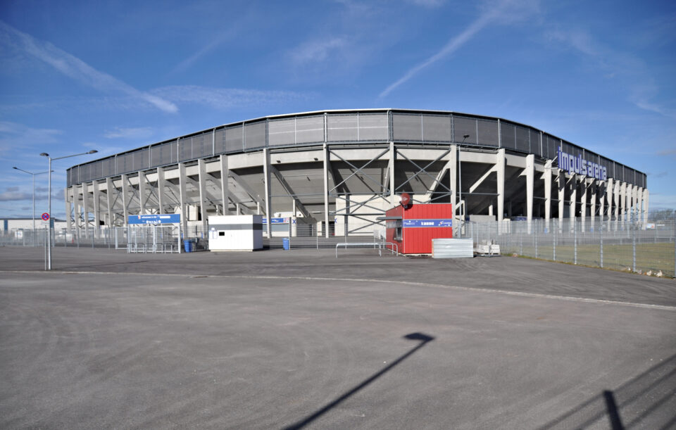 Großes Stadion mit der Aufschrift „impuls arena“ an der Fassade, moderner Bau mit zahlreichen Betonstützen, davor mehrere kleinere Gebäude, Container, Zäune und Lichtmasten, unter klarem Himmel mit Schleierwolken.