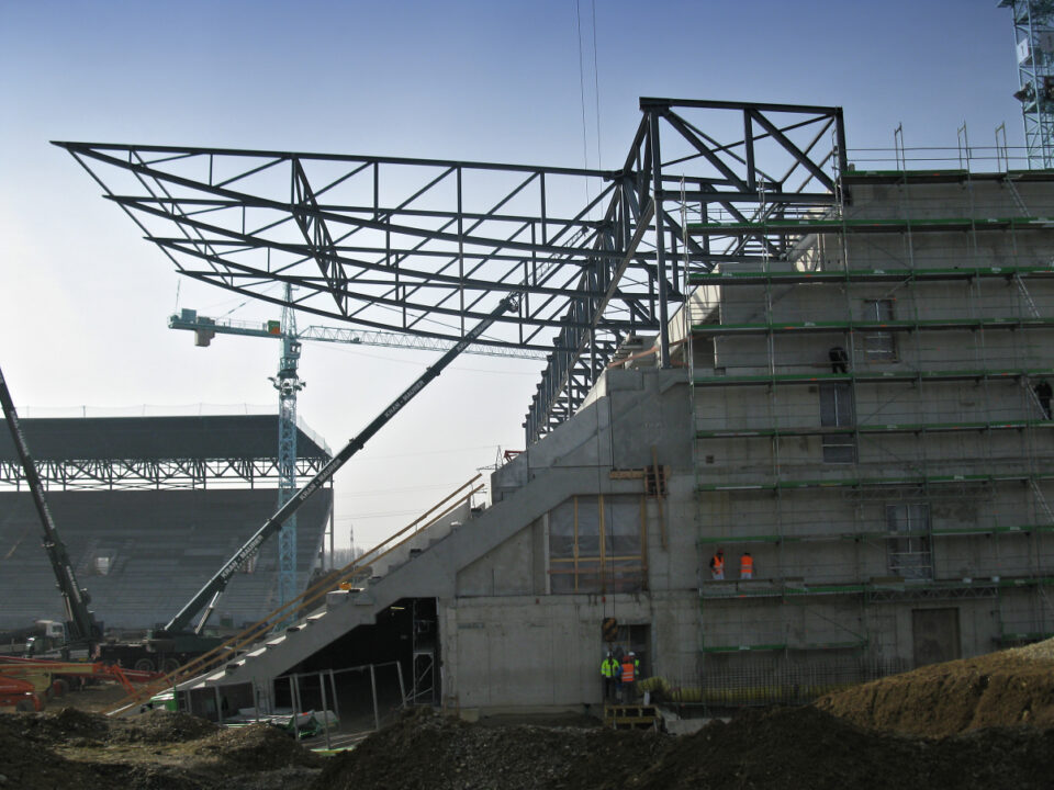 Baustelle eines großen Stadions oder einer Arena, mit sichtbarem Stahlgerüst und Betonteilen, Kränen und Baumaschinen im Hintergrund sowie mehreren Arbeitern auf dem Gelände unter klarem Himmel.