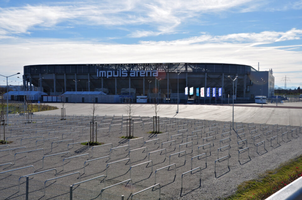 Großes, rundes Stadion mit dem Namen „impuls arena“, davor eine offene Fläche mit mehreren in Reihen angeordneten Metallabsperrungen zur Besucherlenkung, unter klarem Himmel mit einigen Wolken.