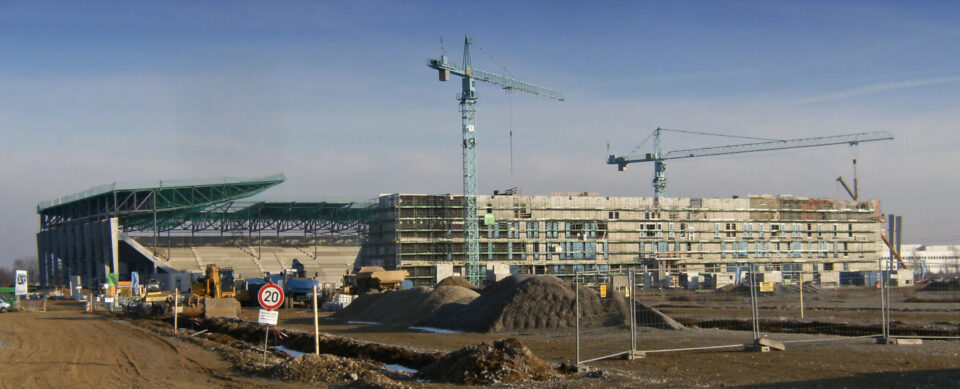 Baustelle mit zwei großen Kränen und mehreren Baufahrzeugen, im Hintergrund ein teilweise errichtetes Gebäude, vermutlich ein Stadion oder Großbau, umgeben von Schotterhaufen, Erdreich und Bauzäunen; links im Bild ein Tempo-20-Verkehrsschild.