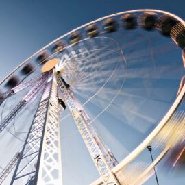 Das Bild zeigt ein großes Riesenrad in Bewegung, aufgenommen aus einem niedrigen Blickwinkel vor klarem, blauem Himmel. Durch die Lichtbeleuchtung und Bewegungsunschärfe der Gondeln entsteht ein dynamischer Eindruck.