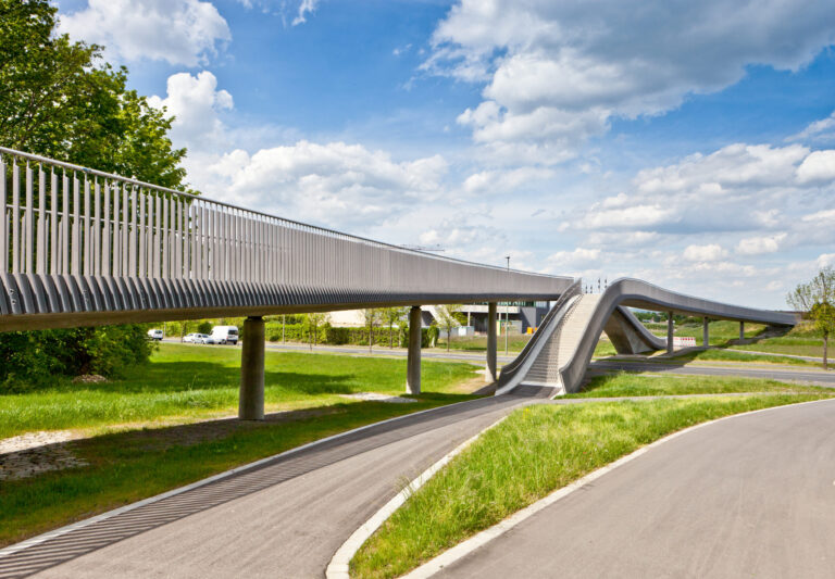 Eine moderne Fußgängerbrücke mit wellenförmigem Design verläuft auf Betonpfeilern über eine Straße, umgeben von grüner Wiese, Bäumen und einem blauen Himmel mit vereinzelten Wolken.