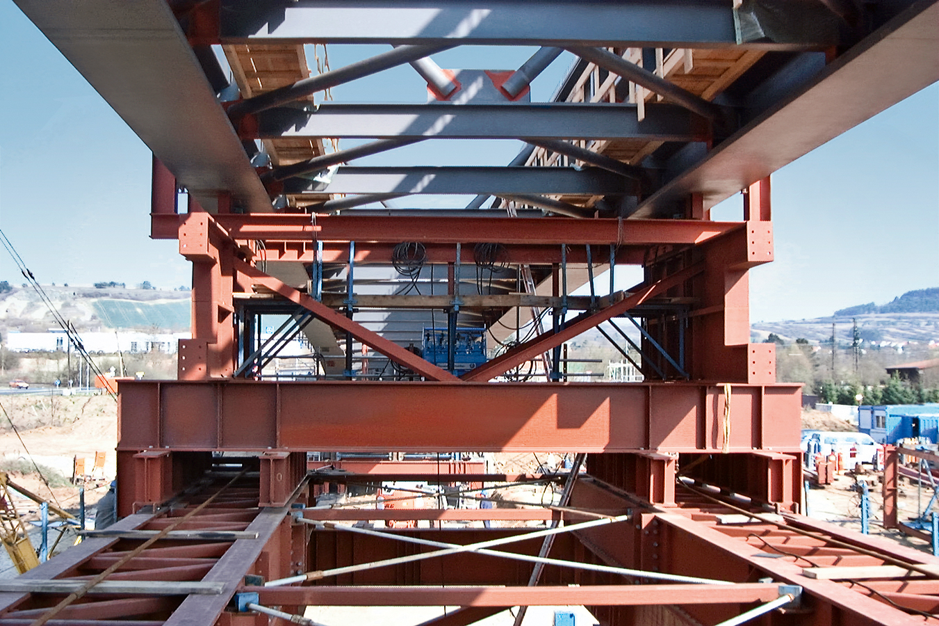 Eine Baustelle mit dem Stahltragwerk eines großen Bauwerks, vermutlich einer Brücke oder eines Gebäudes; sichtbare Gerüste, Baumaterialien und Maschinen, im Hintergrund Hügel und weitere Strukturen.
