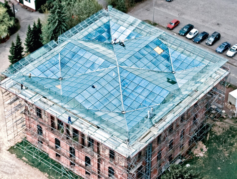 Das Bild zeigt eine Baustelle aus der Vogelperspektive mit einem Gebäude, dessen Dach aus geometrisch angeordneten Glasflächen besteht. Um das Gebäude herum befinden sich Gerüste, und auf dem Dach sind mehrere Bauarbeiter zu sehen. Im Umfeld erkennt man Bäume, parkende Autos und weitere Gebäude.