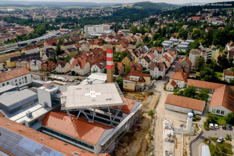 Luftaufnahme einer Stadt mit Wohn- und Industriegebäuden; im Vordergrund ein großes Gebäude mit einem rot markierten Hubschrauberlandeplatz auf dem Dach und ein rot-weiß gestreifter Schornstein, im Hintergrund zahlreiche Häuser mit roten Ziegeldächern, umgeben von Grünflächen und hügeliger Landschaft.