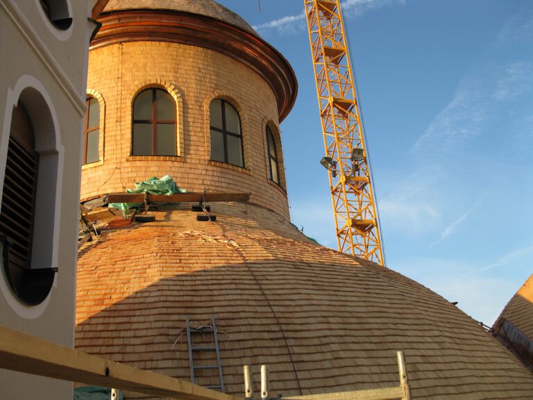 Baustelle mit großer, kuppelförmiger Dachkonstruktion aus Holzschindeln, daneben ein gelber Baukran; auf dem Dach befinden sich grüne Planen und Baumaterialien, sowie eine Leiter, das Gebäude zeigt dunkle Rundbogenfenster unter klarem Himmel.