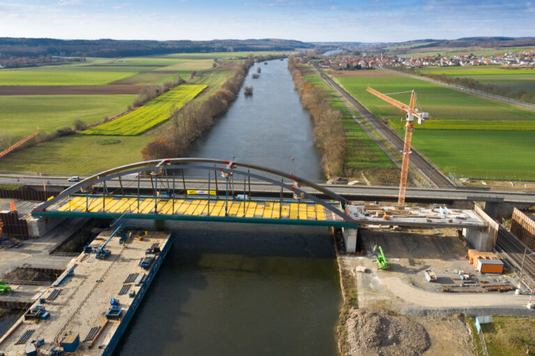 Im Bau befindliche Bogenbrücke über einen Fluss mit gelben Arbeitsplattformen auf der Oberseite; rechts steht ein Kran, im Hintergrund sind Felder, Straßen und eine kleine Ortschaft zu sehen.