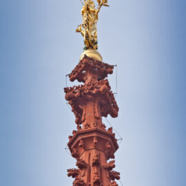 Das Bild zeigt die Spitze eines hohen, kunstvoll gestalteten Bauwerks aus rötlich-braunem Material mit filigranen Verzierungen und einer goldenen Statue einer Figur mit einem Gegenstand in der Hand vor klarem, blauem Himmel.