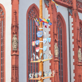 Das Bild zeigt einen hohen, bunt geschmückten Holzpfahl mit Wappen, Fahnen und Figuren vor einem historischen Gebäude mit rot-weißer Fassade und verzierten architektonischen Details.