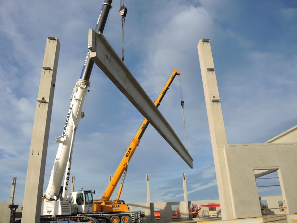 Baustelle mit zwei Kränen, die einen großen Betonträger zwischen mehreren hohen Betonsäulen einsetzen, unter klarem Himmel mit einigen Wolken.