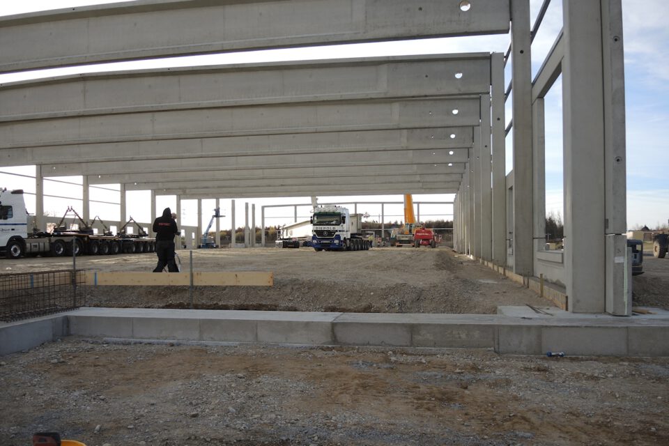 Baustelle mit einem teilweise errichteten Gebäude aus großen Betonträgern und -säulen, mehreren Fahrzeugen wie einem Lkw und Baumaschinen sowie einer Person im Vordergrund, die offenbar am Bau beteiligt ist; der Boden ist mit Kies und Erde bedeckt.