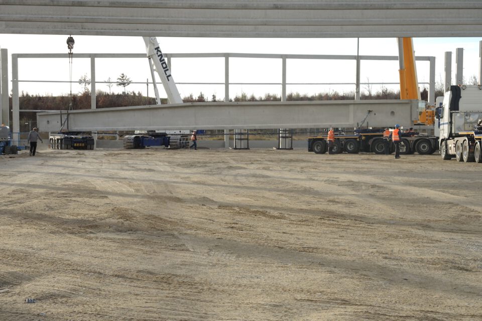 Baustelle, auf der ein großer Betonträger mithilfe von Kränen angehoben wird, mit mehreren Arbeitern in Warnkleidung und Schutzhelmen sowie schwerem Gerät wie Kränen und Lkw auf einem erdigen Untergrund.